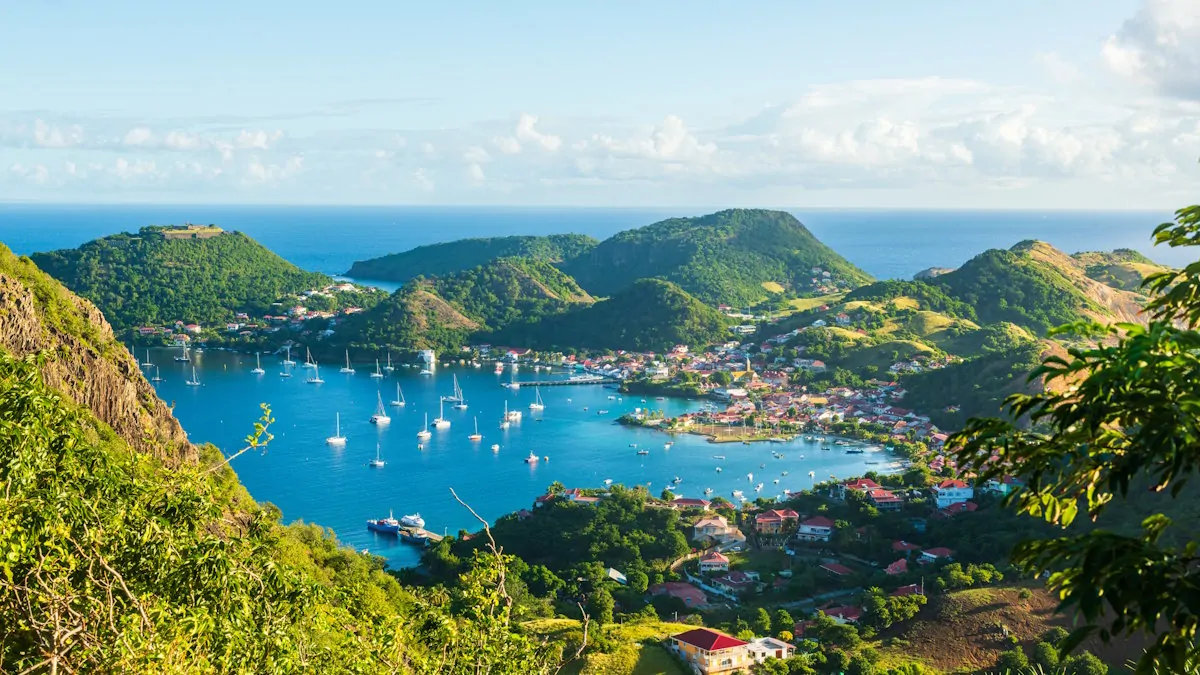 Vue panoramique sur une baie tropicale entourée de collines verdoyantes, avec un village côtier et de nombreux voiliers au mouillage.